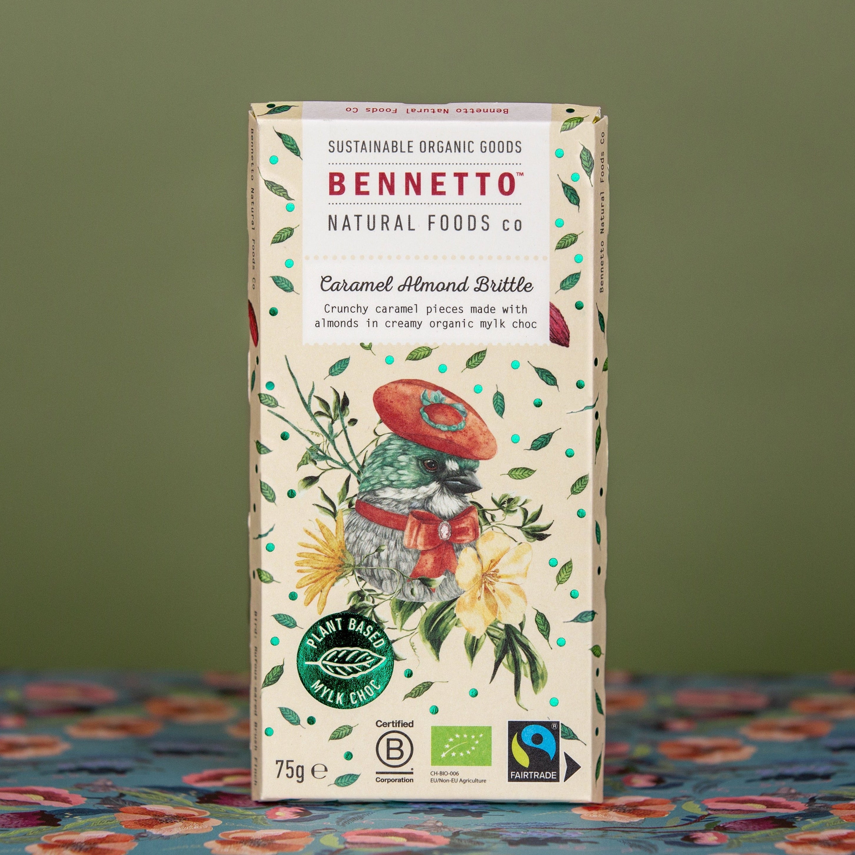 A bar of Bennetto Natural Foods Co Caramel Almond Bruittle Mylk chocolate bar. Featuring a light yellow tone bar background colour with green leaves, and a bird illustration, with dark green background and floral blue and red tablecloth.