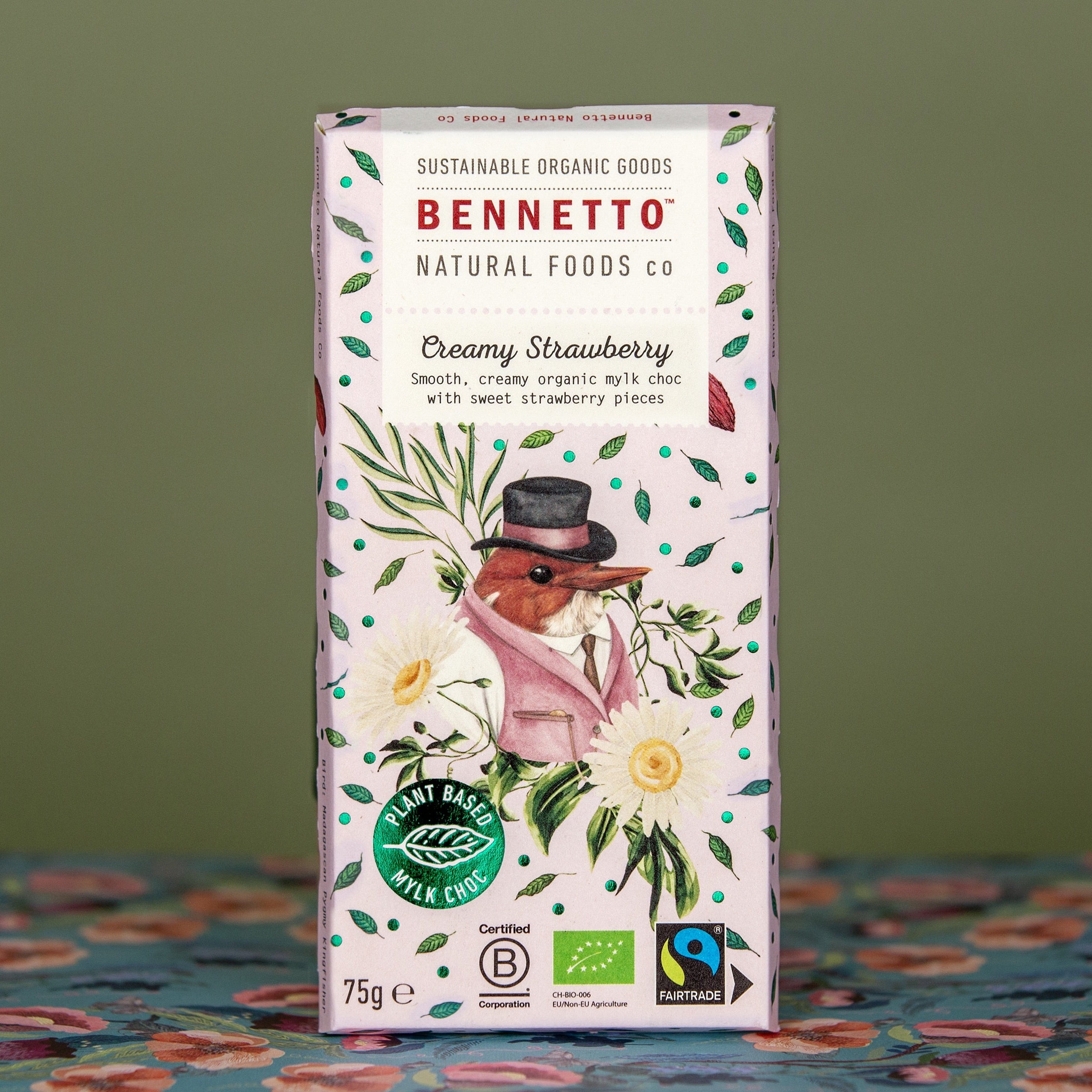 A bar of Bennetto Natural Foods Co Creamy Strawberry MYLK chocolate. Featuring a light pink bar background with green leaves and a pink dressed bird illustration. With a dark green blurred background and blue and red tablecloth.
