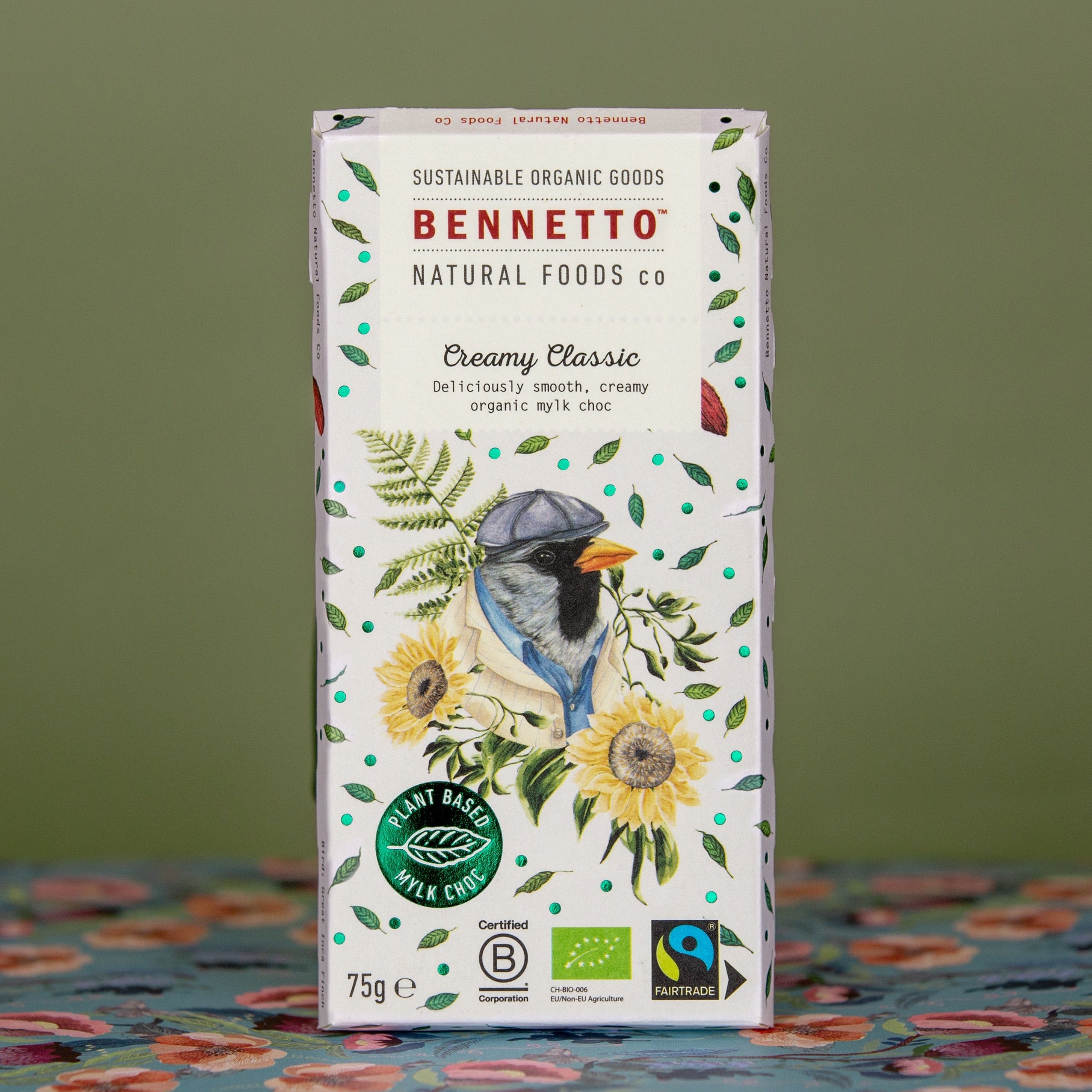 A bar of Bennetto Natural Foods Co. Creamy Classic MYLK Chocolate, featuring a light blue bar background and a bar with green leaves, yellow sunflowers, and a blue bird illustration, standing on a patterned blue tablecloth against a muted green background.