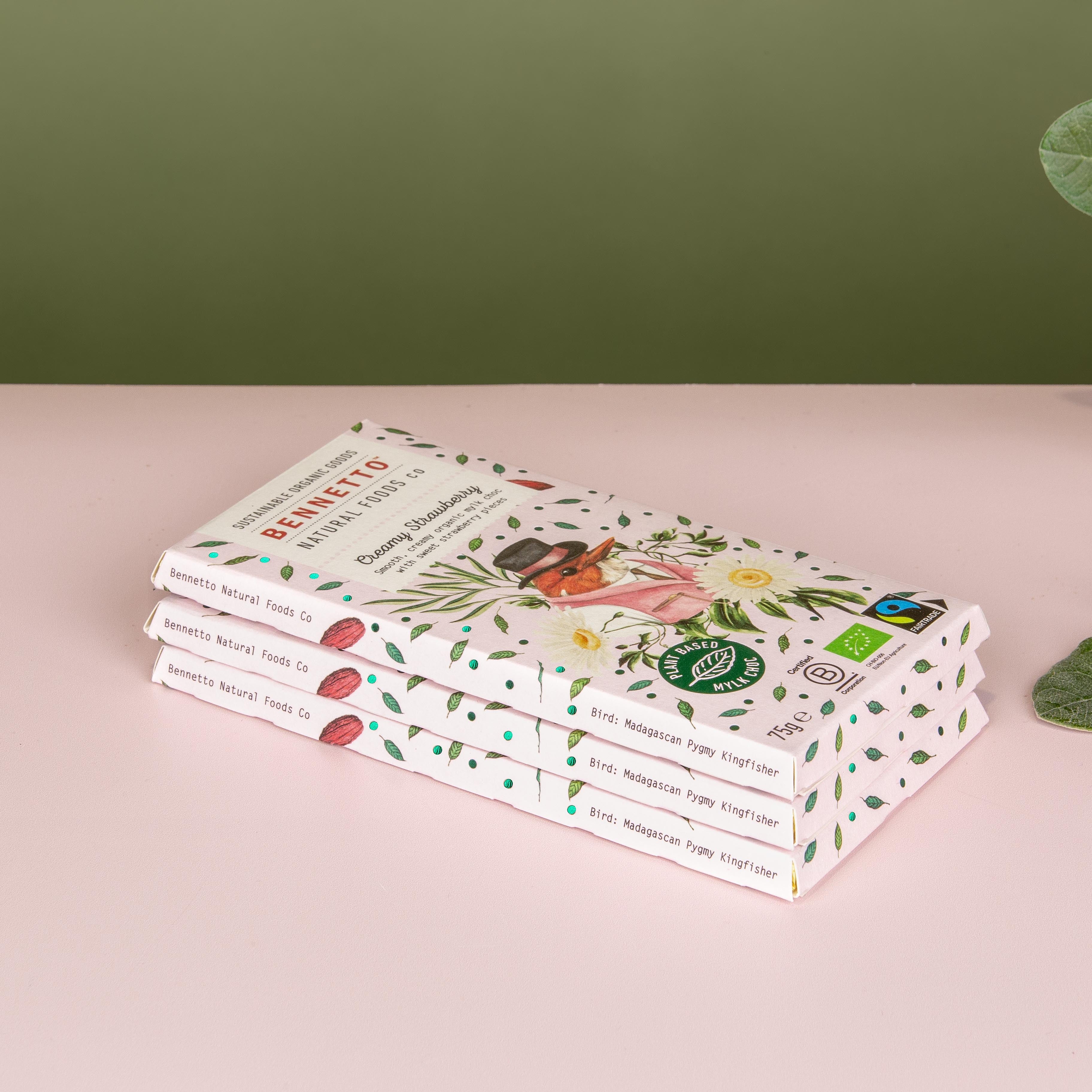 Three stacked boxes of Bennetto Organic chocolate bars, decorated with botanical illustrations, set on a light pink surface against a muted green background with eucalyptus leaves on the right.