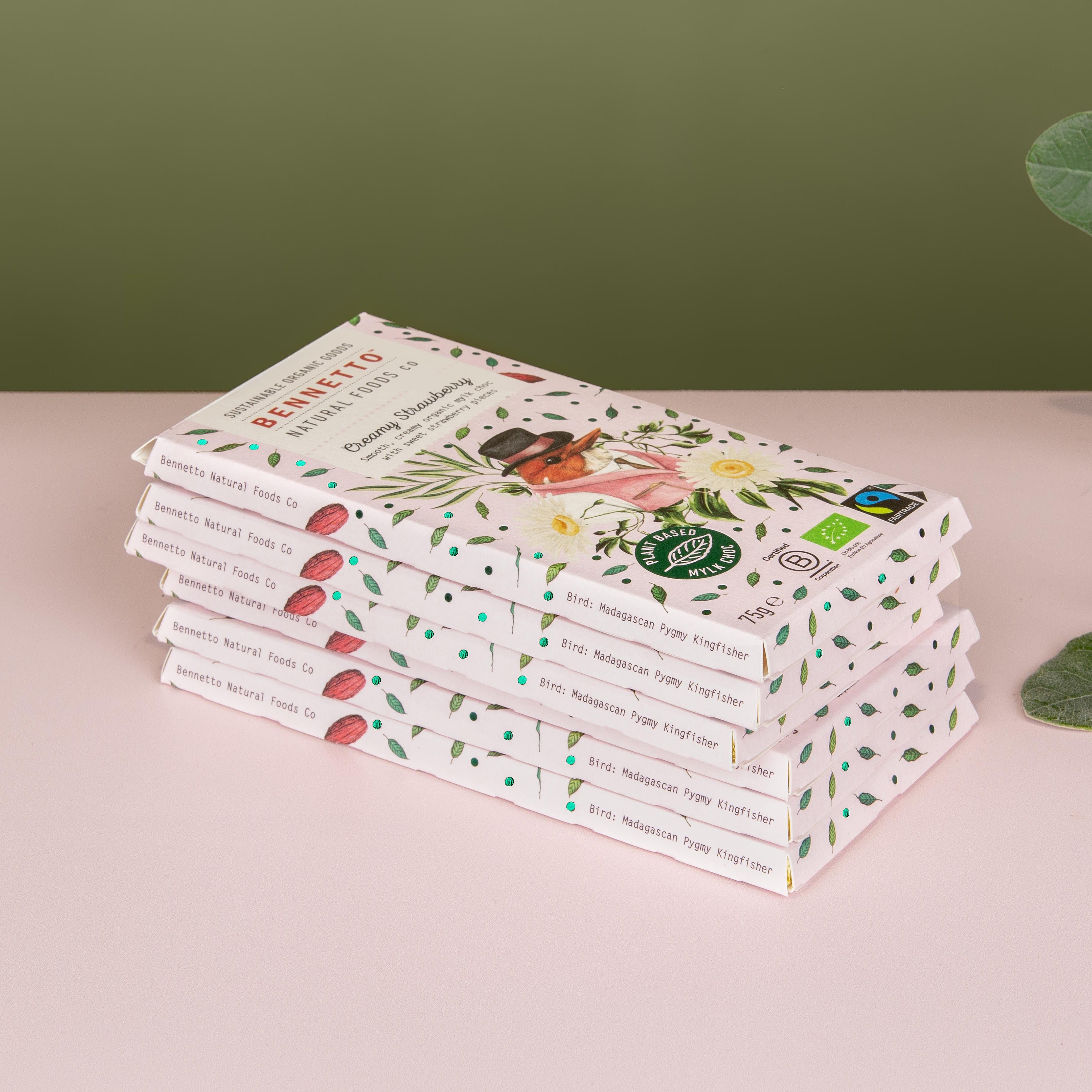 A stack of mylk chocolate bars in decorative packaging, placed on a pale pink surface against an olive green background, with eucalyptus leaves on the right side.