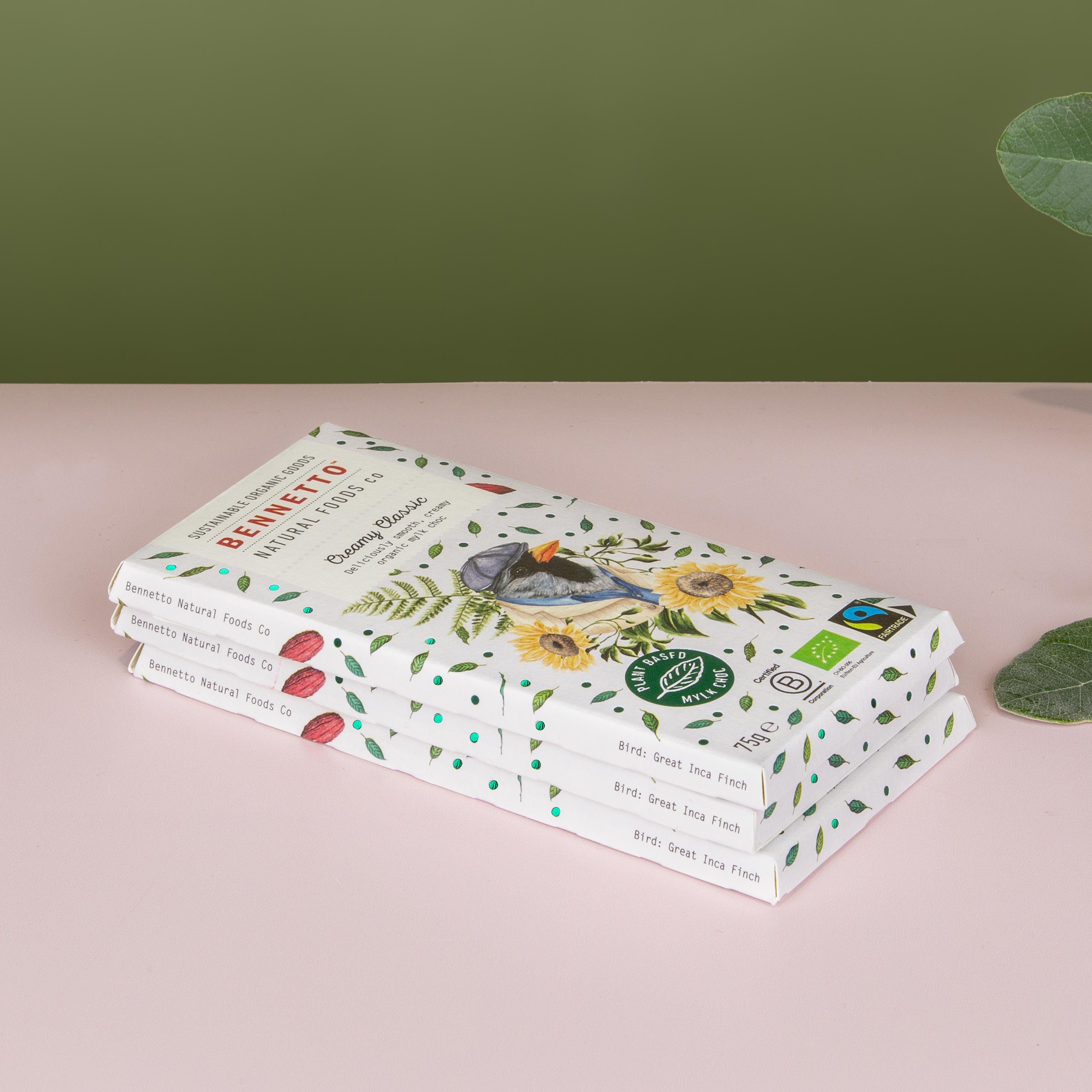 A stack of Bennetto organic dark chocolate bars in light blue packaging decorated with green leaves and yellow sunflowers, set on a pink surface against an olive green background with eucalyptus leaves on the right.