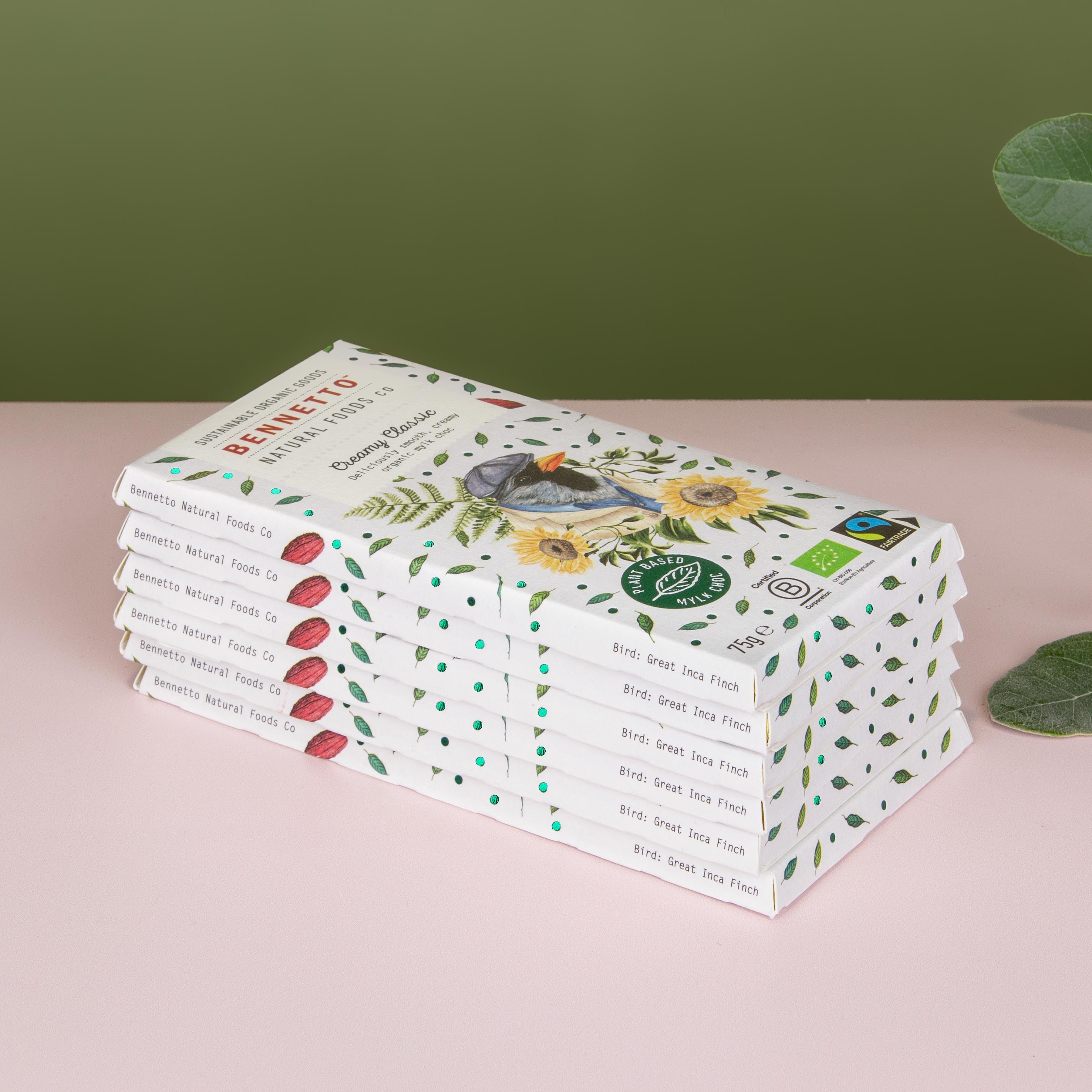 A stack of Bennetto organic chocolate bars on a pink surface with a green background and eucalyptus leaves on the right.