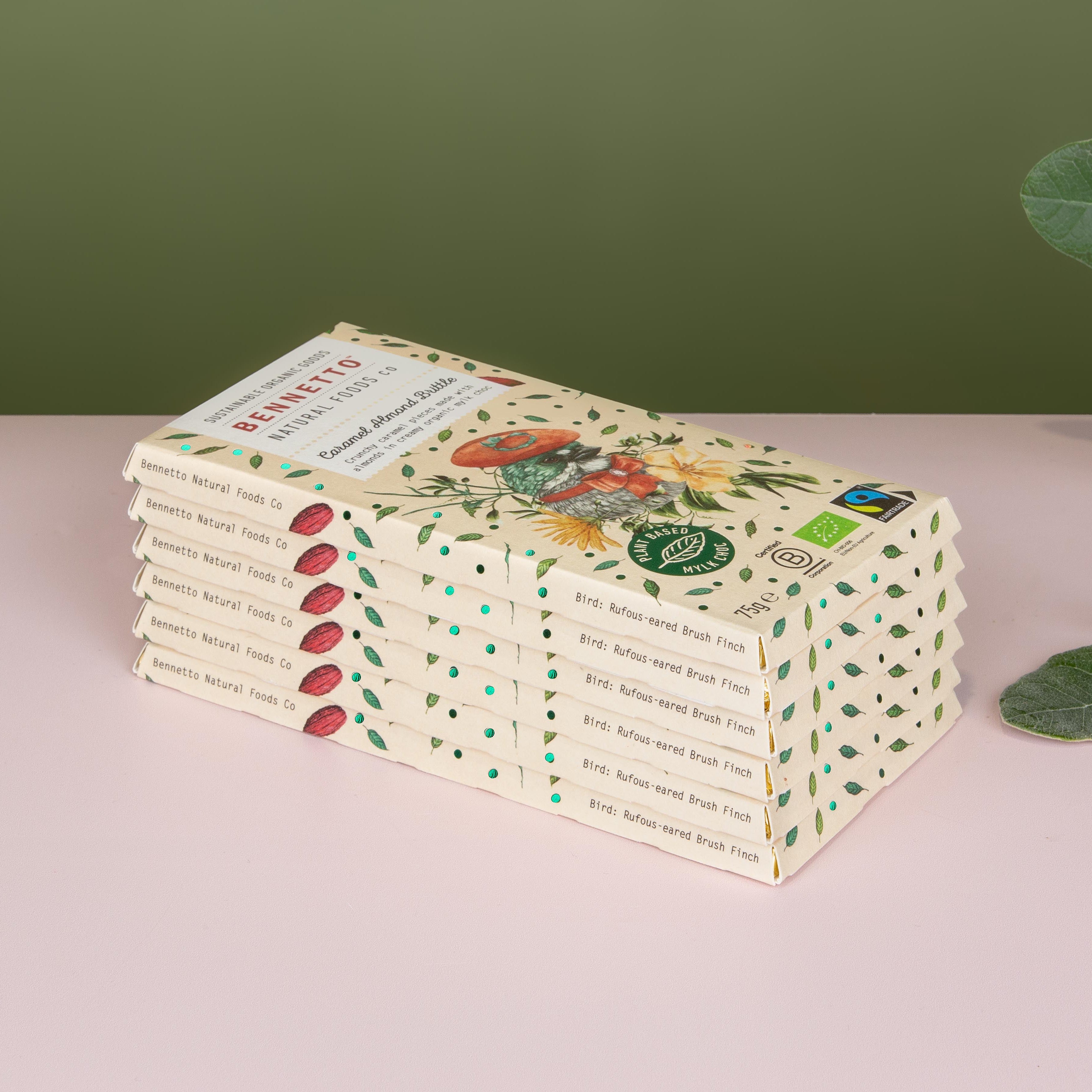 A stack of Bennetto organic chocolate bars in cream-colored packaging with green leaf patterns, set on a light pink surface against a dark olive green background with eucalyptus leaves on the right.