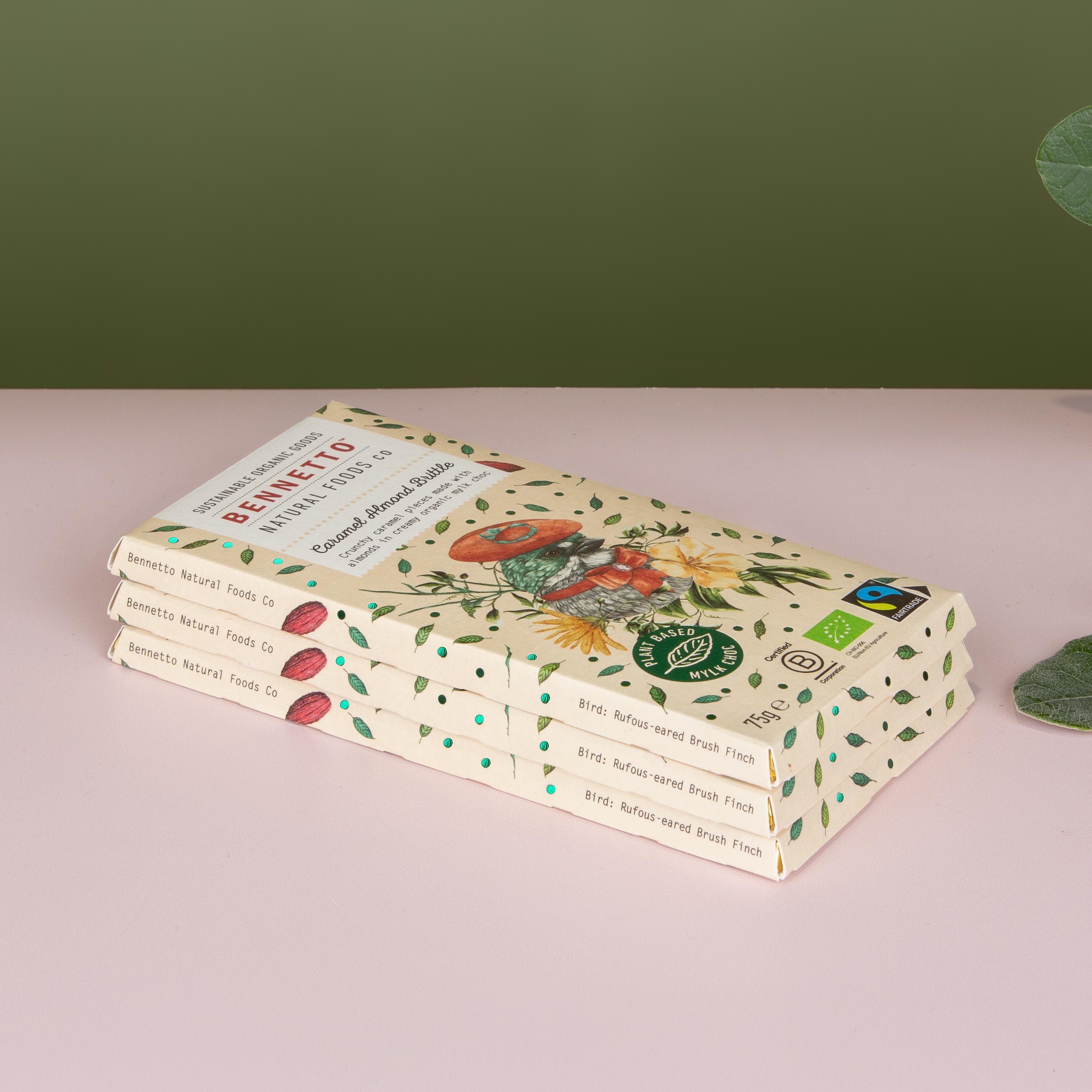 A stack of three Bennetto chocolate bars in their illustrated paper packaging, placed on a light pink surface against a dark olive green background with eucalyptus leaves on the right.