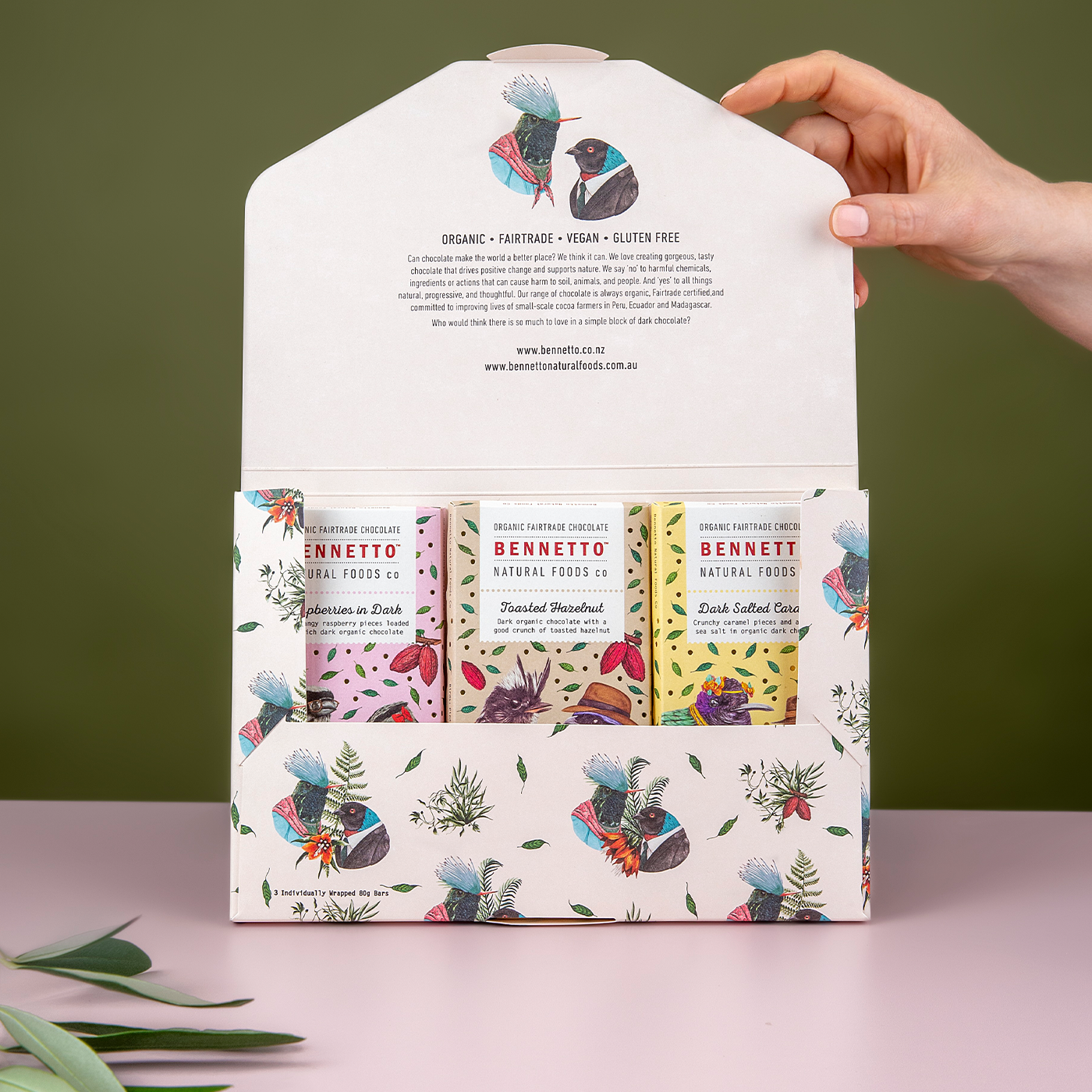 A hand holding an open gift box of Bennetto chocolate bars with three different flavors displayed: Raspberry in Dark, Dark Salted Caramel, and Toasted Hazelnuts.