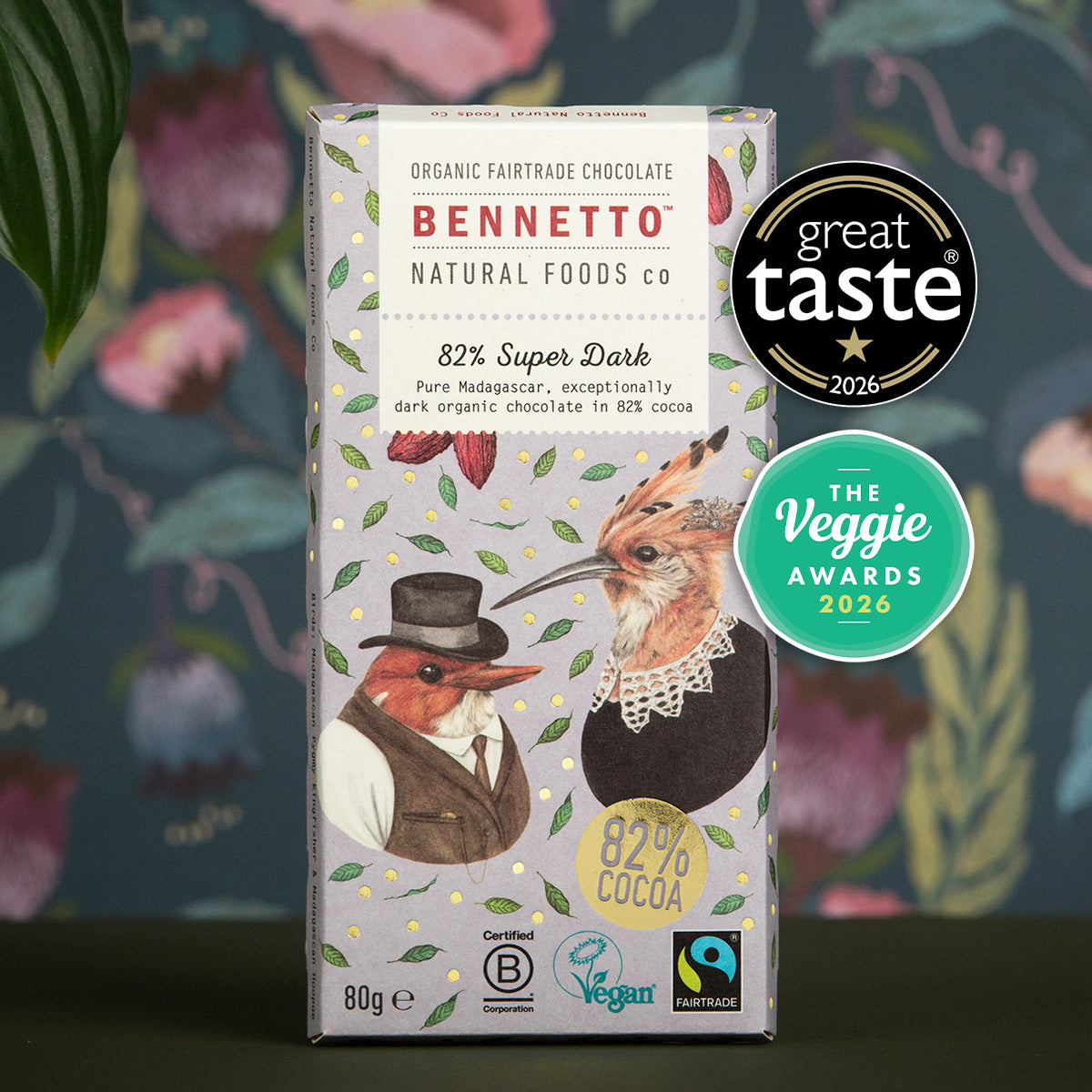 A boxed bar of Bennetto Natural Foods Co chocolate stands upright against a decorative floral background. The package is white with red, green, and brown illustrations and award badges on the right side.